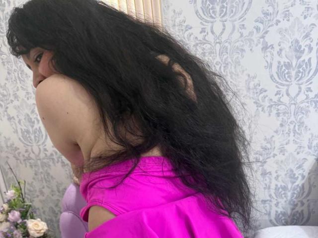 Diana Lane, com cabelo preto longo, blusa rosa e saia rosa, agachada, mostrando suas costas e nádegas, olhando por cima do ombro com um sorriso tímido..