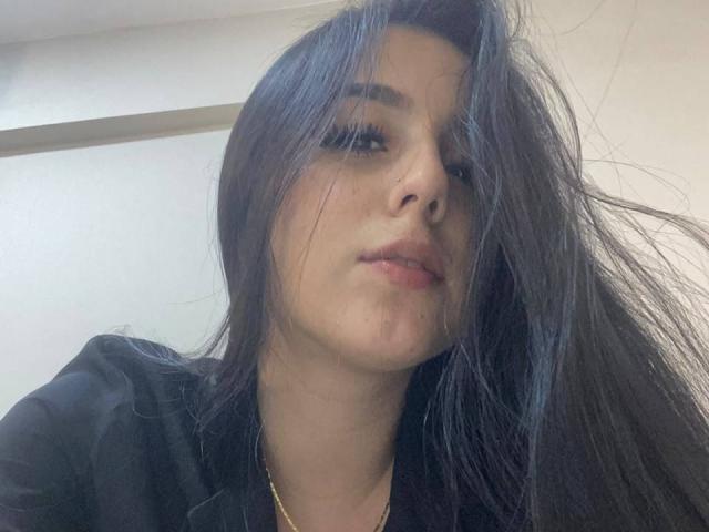 StefanyDelly has long, messy black hair covering her eyes, a nose piercing, and a slight smirk with pink lips, wearing a black shirt and gold necklace.