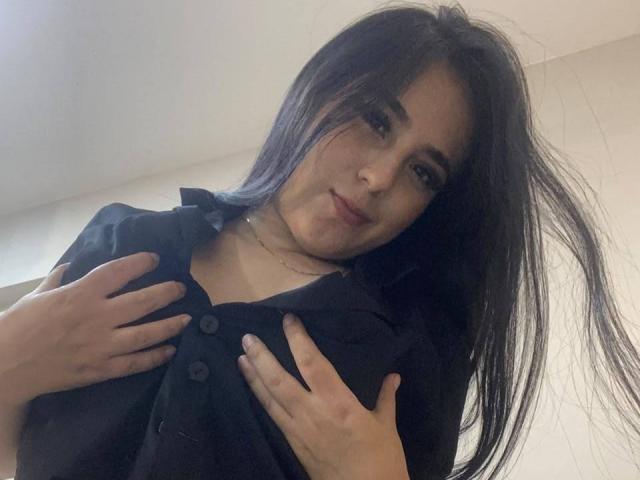 StefanyDelly with long black hair, wearing a black shirt, hands on her tits, slightly smiling, low-angle shot, soft lighting, casual look, slightly messy hair.