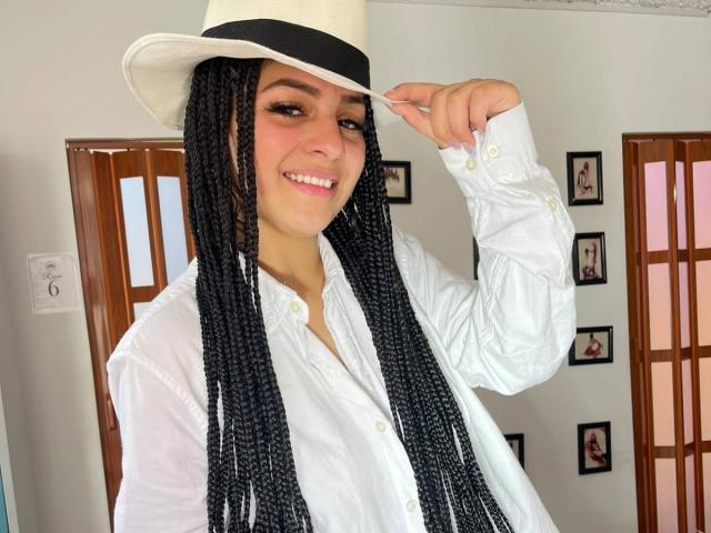 LonxEva with long black braids, wearing a white hat and shirt, smiling at the camera, holding the hat with her right hand, showing her cute face and tits.
