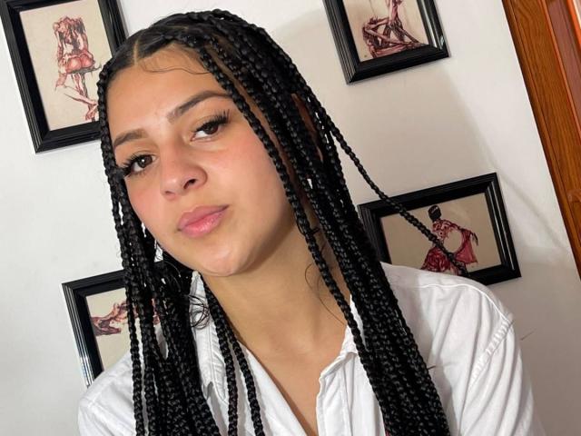 LonxEva with long black braids, light brown skin, and pink lips, wearing a white shirt, has a relaxed, slightly smirking face, brown eyes, and subtle makeup.