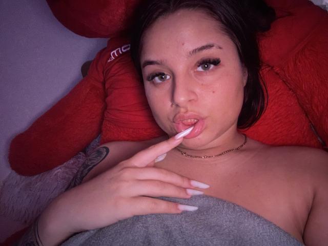 Lucie, de pelo rojo, con uñas largas, labios ligeramente entreabiertos y un dedo en la boca, sin ropa, mostrando un tatuaje en su brazo, acostada contra un peluche rojo, y con un collar de cadena puesto..