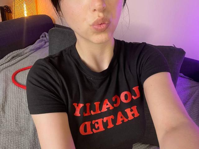 AmaiRin met volle lippen, een zwart T-shirt met rode tekst, kort donker haar, bleke huid en kleine borsten, zittend op een grijze, gestructureerde bank..