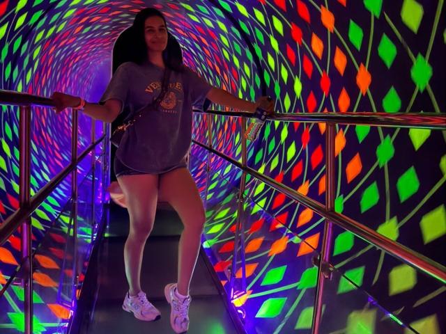 AmberGray69, con cabello negro largo, vestida con una camiseta gris y zapatillas blancas, está de pie en una escalera colorida e iluminada con luces de neón, con las manos sobre los pasamanos de metal y las piernas separadas..