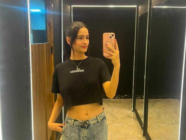 Melody Mansson, con i capelli scuri, un top corto nero che lascia scoperto l'addome, e jeans blu, sta scattando un selfie allo specchio, tenendo in mano un telefono rosa, indossando una collana e sorridendo leggermente..