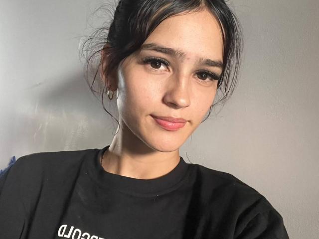 MelodyMansson with dark hair in a messy bun, wearing a black shirt, has brown eyes, full pink lips, and small earrings, looking at the camera with a slight smile.