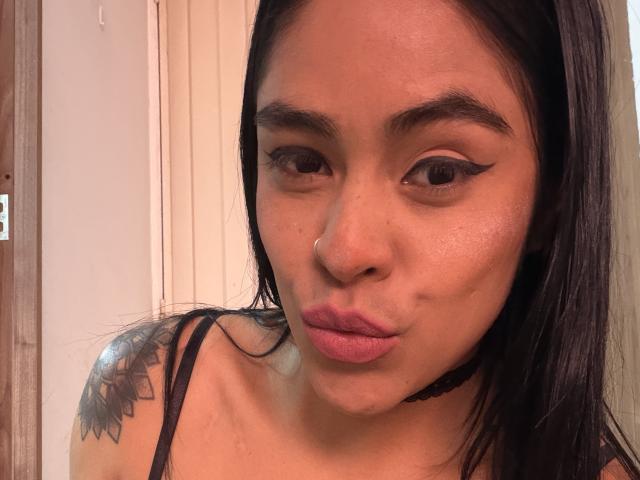 StellaBeckker with puffy lips, black eyeliner wings, nose ring, black choker, black hair, tattoo on right shoulder, wet skin, wearing black strap, looking at camera.