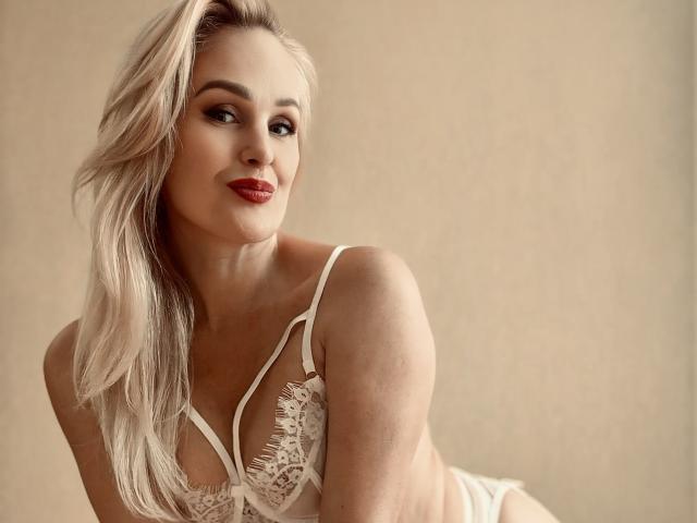 MiaDreamy, con i capelli biondi, il rossetto rosso e il reggiseno di pizzo bianco, mostra il seno e sorride. Luce soffusa, posa delicata..