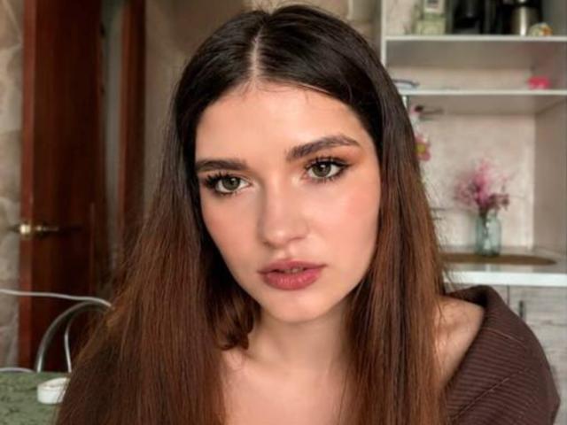 Elenore Harajly tem cabelos castanhos longos, olhos verdes e lábios rosados cheios. Ela está vestindo uma blusa marrom com um ombro de fora e tem um leve rubor nas bochechas. Seus seios não estão visíveis..
