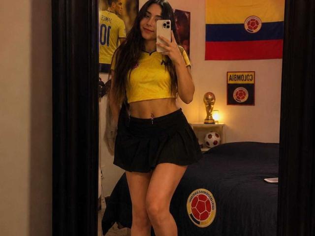 MindyLove con lunghi capelli castani, top giallo, minigonna nera, che tiene in mano un telefono, selfie allo specchio, che mostra il ventre, i glutei e le gambe, in una camera da letto con decorazioni a tema calcistico..