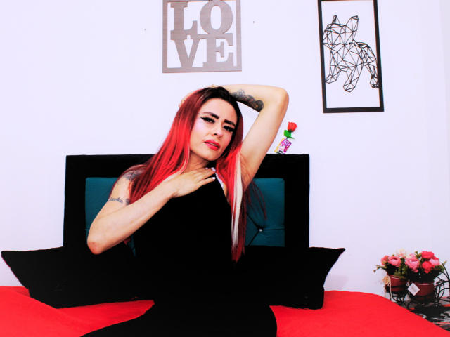 VogueB avec des cheveux roux et noirs, une robe noire sans manches, un tatouage sur le bras, une main dans les cheveux, assise sur un lit rouge, avec des oreillers noirs, un mur blanc, des œuvres d'art en noir et blanc, un vase à fleurs..