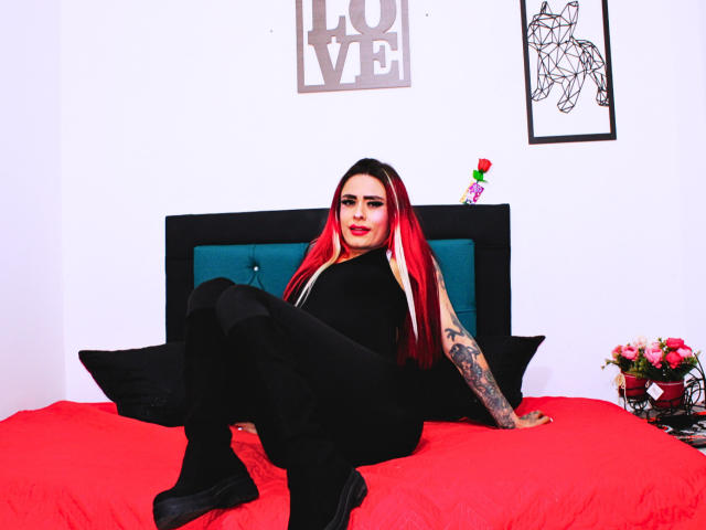 VogueB com cabelo ruivo e preto, blusa preta sem mangas, calças pretas e botas pretas, sentada em uma cama vermelha, mostrando um braço tatuado e uma rosa charmosa..