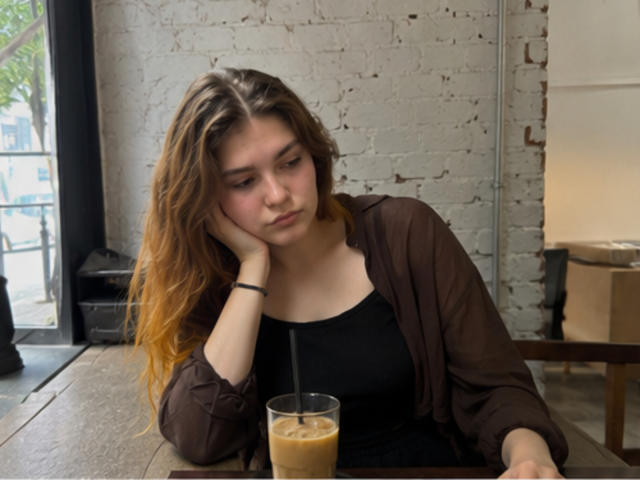 ElenoreHarajly with wavy brown hair, wearing a brown cardigan and black top, sipping iced coffee, resting head on hand, looking down, sitting at a wooden table.