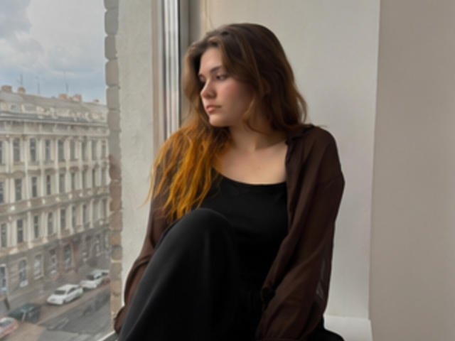 ElenoreHarajly with long brown hair and yellow tips, sitting by a window, wearing a black dress and brown cardigan, looking outside with a serious expression.