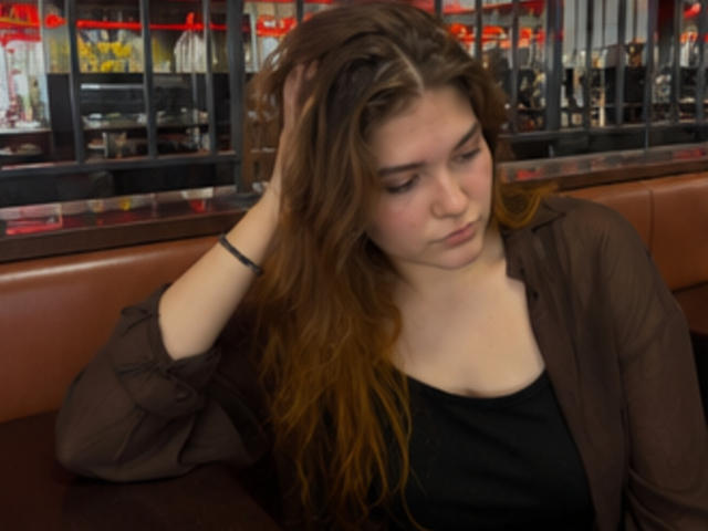 Elenore Harajly, com cabelos castanhos longos, olhos castanhos e pele clara, vestindo uma blusa preta e uma camisa marrom, sentada em um assento de couro vermelho, com a mão no cabelo, olhando para baixo..