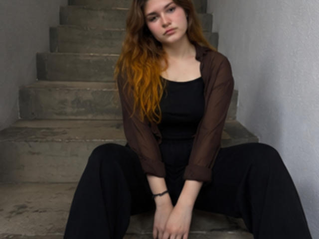 Elenore Harajly, con el pelo largo y castaño, está sentada en unos escalones de hormigón, vestida con un vestido negro y una blusa marrón transparente, con las piernas separadas, luciendo cansada y aburrida, y con una pulsera visible en la muñeca..