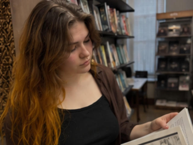 Elenore Harajly heeft lang, golvend bruin haar met oranje punten, draagt een zwarte top en een bruin vest, en leest een krant, waarbij ze naar beneden kijkt met een subtiele, geconcentreerde uitdrukking..