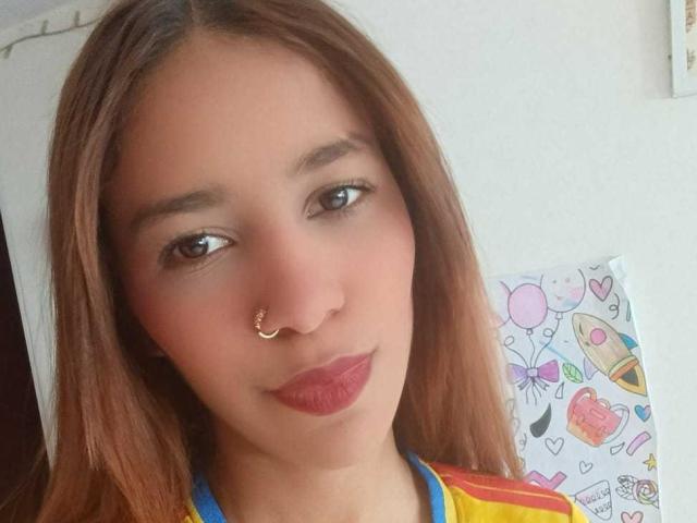 ValerieWoods has light brown skin, long brown hair, brown eyes, red lipstick, and a small nose ring. She's wearing a colorful top, looking at the camera with a slight smile.