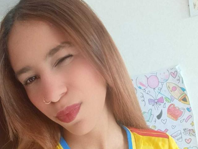 ValerieWoods has light brown skin, winking with one eye, nose ring, red lipstick, and long brown hair, wearing a yellow and blue shirt, showing a cute smile.
