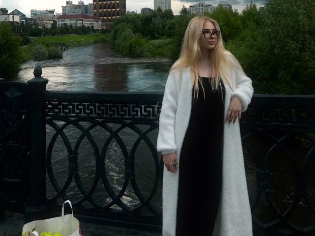 Chassidy Kulju, met lang blond haar, een bril, een zwarte jurk en een witte bontjas, staat op een brug, met een tas in haar hand, aan de rivier, op een bewolkte dag..