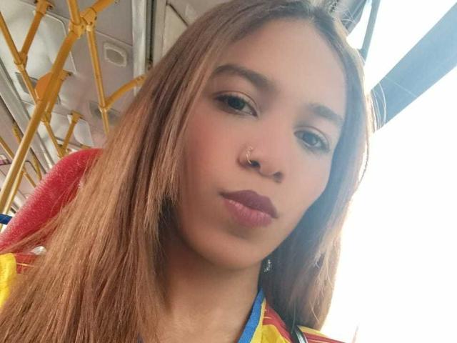 ValerieWoods has brown hair, a nose ring, and pouty lips with pink lipstick. She's wearing a colorful top and sitting on a yellow bus.