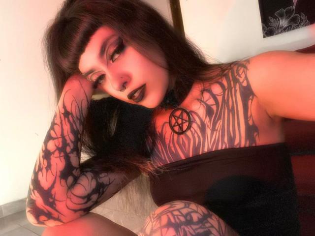 LitenKiitt trägt dunkelroten Lippenstift, schwarzen Eyeliner und einen Nasenring, hat Tattoos, die ihre Brust und ihren Arm bedecken, trägt ein schwarzes Oberteil und sitzt mit einer Hand auf dem Kopf..