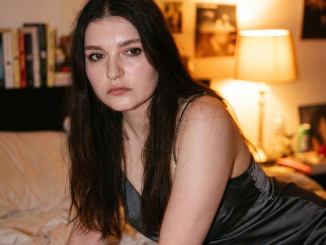 Elenore Harajly tiene el cabello largo y oscuro, una expresión seria, y lleva un vestido negro de seda, inclinándose hacia adelante sobre una cama con una lámpara que emite una luz cálida en el fondo..