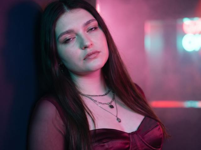 Elenore Harajly hat langes, braunes Haar, helle Haut und trägt ein rotes, seidenes Kleid mit Ausschnitt und mehrere Halsketten. Sie blickt leicht gelangweilt nach unten..