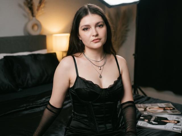 Elenore Harajly, à la peau pâle et aux longs cheveux noirs, portant de la lingerie en dentelle noire, un corset et des manches en mesh noir, assise sur un lit noir, regardant la caméra, avec un sourire discret..