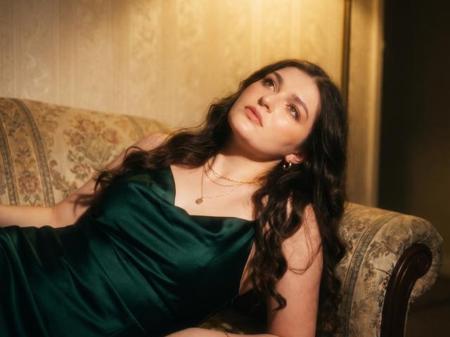 Elenore Harajly, met lang bruin haar, een groene satijnen jurk, een gouden ketting en gouden oorbellen, leunt achterover op een sofa met een bloemenprint, en kijkt op met een ontspannen, licht verleidelijke uitdrukking..