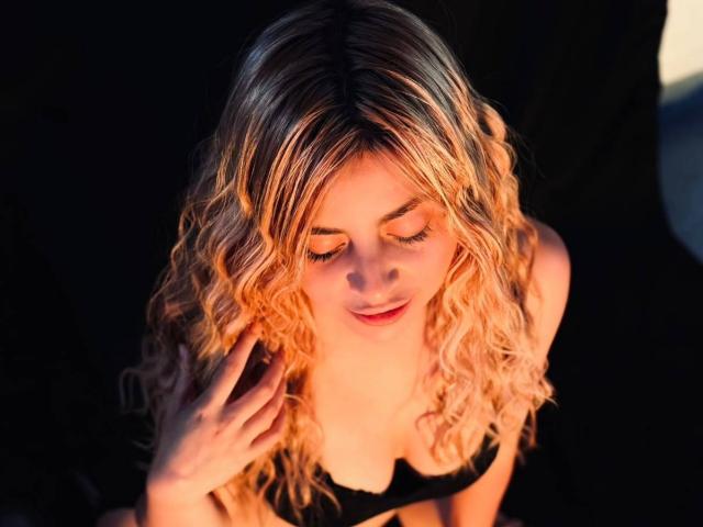 Celeste, traviesa, con cabello rubio rizado, ojos cerrados, sostén negro, mano cerca del rostro, luz naranja suave, ligera sonrisa, expresión suave, sombras delicadas, tono de piel cálido, pose relajada..