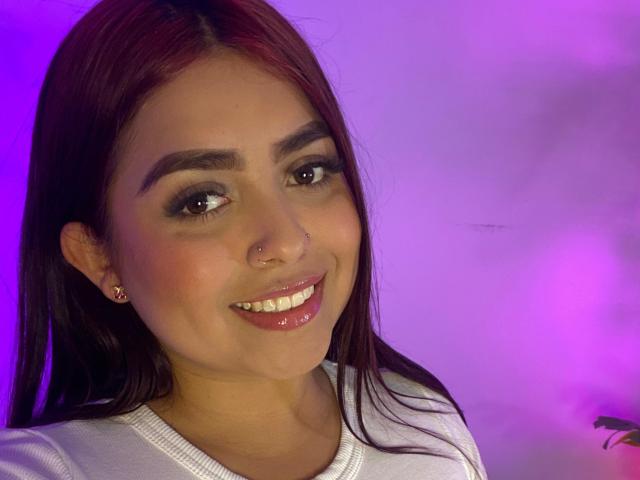 SammyOwenss tem cabelo ruivo, olhos castanhos, um piercing no nariz e está sorrindo, com lábios rosados e brilhantes. Ela está vestindo uma camisa branca, mostrando seus dentes brilhantes..