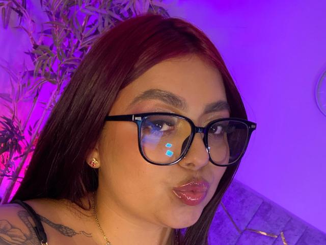 SammyOwenss tiene el cabello rojo oscuro, gafas negras, labios rosados brillantes y un tatuaje en el hombro. En el fondo, hay luces moradas y rosas..