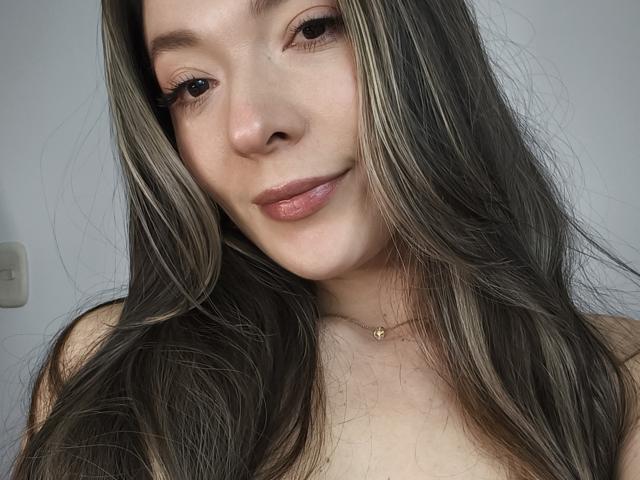 TaniaSandersX, com cabelos longos e castanho-escuros, pele clara e batom rosa, usando um colar de ouro pequeno, exibindo seus seios em uma selfie..