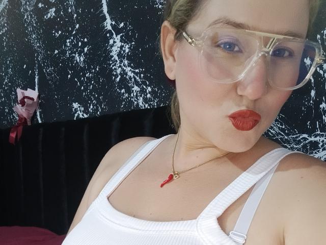 AlexaHotSeins with glasses, pouty red lips, white tank top, necklace with red gem, fair skin, blonde hair, pinkish eyeshadow, slightly chubby, black and white textured wall background.
