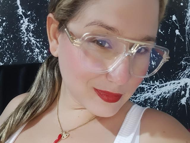 AlexaHotSeins with red lipstick, clear glasses, and a white tank top, showing her face and chest. She has a gold necklace with a red pendant. Blonde ponytail.