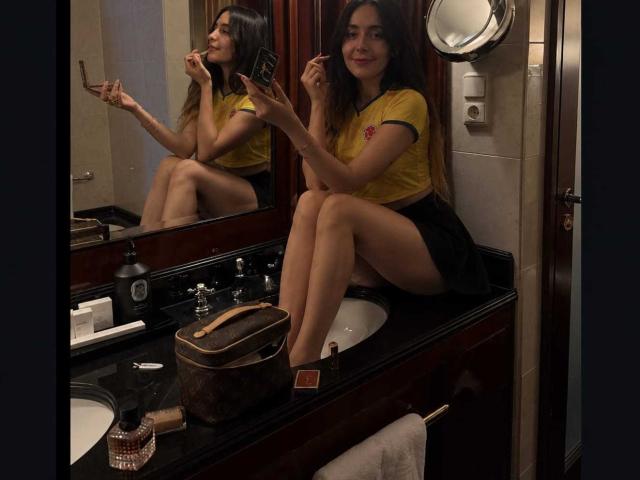 MindyLove with long brown hair, yellow shirt, and black skirt, sitting on a bathroom counter, taking a selfie with her phone, showing her bare ass in the mirror.
