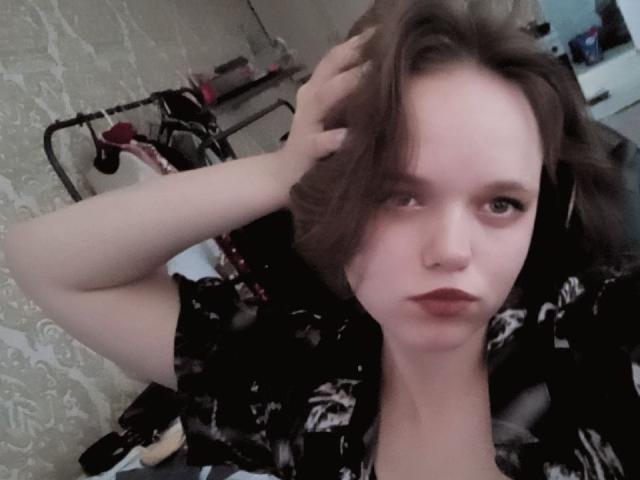AngeliaAmstutz with short brown hair, pouty lips, and wearing a black floral shirt, has one hand in her hair, looking at the camera, pale skin, and a nose ring.