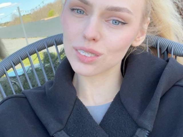 Margorie Symon hat eine helle Haut, blaue Augen und zeigt ihre Zähne mit einem leichten Lächeln. Sie trägt einen schwarzen Mantel, und ihr blondes Haar ist zu einem Pferdeschwanz gebunden..