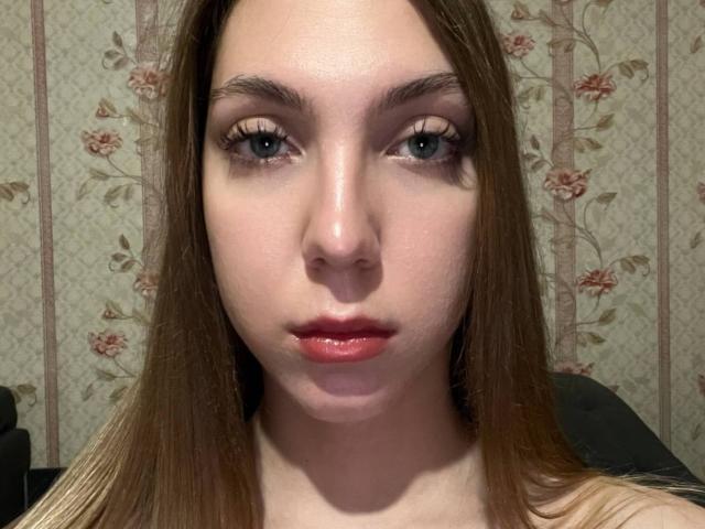 FarahSings tiene la piel clara, cabello castaño lacio, ojos azules y lápiz labial rojo. Sus cejas son oscuras y está mirando directamente a la cámara con una expresión neutra..