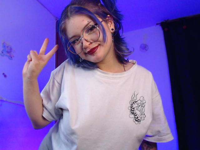 RavenThuwu, con i capelli blu, gli occhiali e il rossetto rosso, indossa una camicia bianca con un disegno di un fiore nero, mostra il segno della pace e lascia intravedere il seno. La stanza è illuminata di blu..