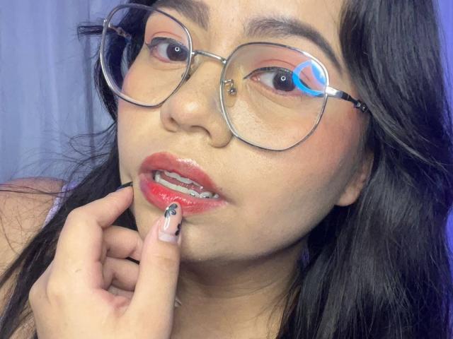 LilyRousee heeft glanzende rode lippen, een neuspiercing en ze bijt in haar vinger met gelakte nagels. Ze draagt ronde brillen en heeft donker, golvend haar..