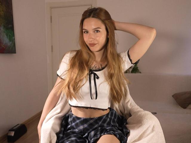 VegasV a de longs cheveux blonds, de petites seins, porte un crop top blanc avec des détails noirs et un short à carreaux, et est assise sur un canapé beige, une main dans ses cheveux..