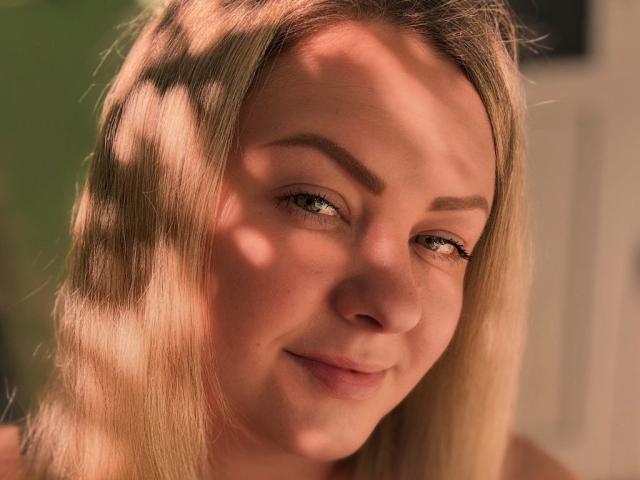 DanielaFloral tiene cabello rubio, ojos verdes y una sonrisa sutil, con ligeras sombras en su rostro. Sus cejas son oscuras y está mirando ligeramente hacia la derecha..