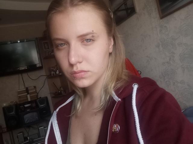 VikiCharm with blonde hair, piercing blue eyes, and a nose ring, wearing a maroon hoodie with white straps, showing her pierced lower lip and small tits.