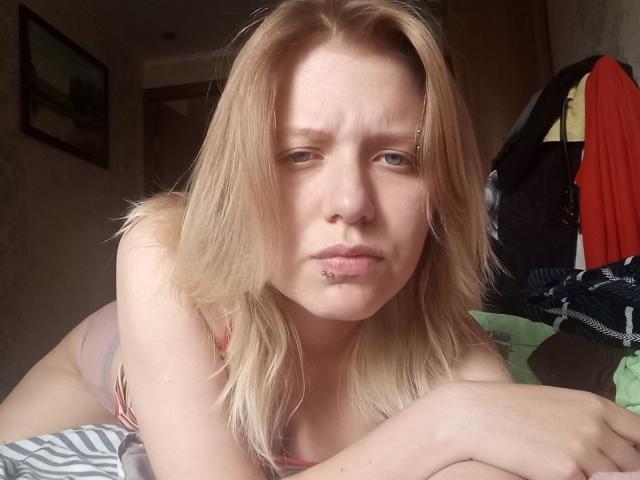 VikiCharm, con capelli biondi, piercing al naso e al labbro inferiore, indossa biancheria intima rosa, ha un'espressione seria, le mani sono sul letto, la stanza è disordinata con vestiti sullo sfondo..