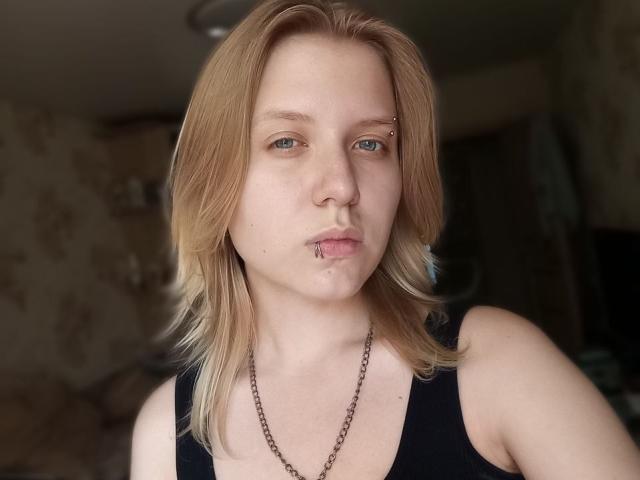 VikiCharm tem cabelo loiro, um piercing no nariz e um piercing no lábio. Ela está vestindo uma camiseta preta com um colar de corrente, e seus olhos azuis estão olhando para a câmera..