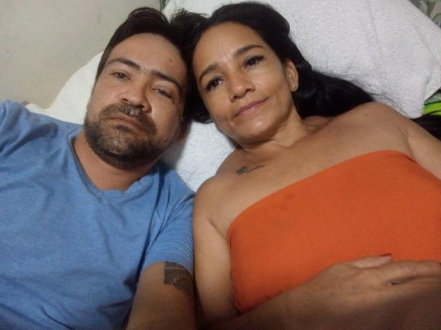 Amantess with medium brown skin, black hair, and small tits, lying topless in orange, beside bearded guy in blue shirt, both on white pillow, smiling at camera.