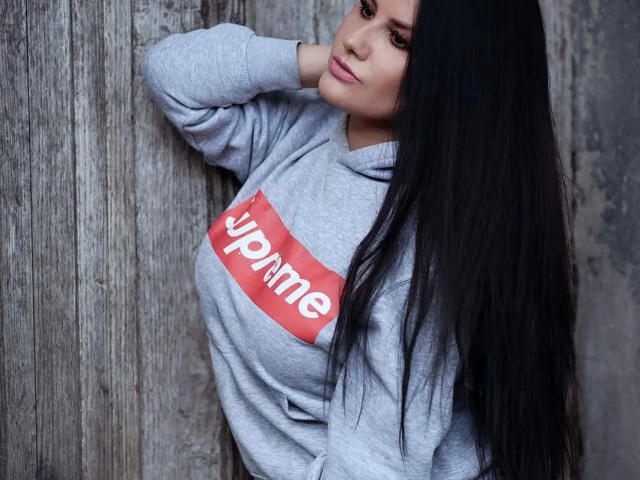 Sydney-Pearl-belle, avec de longs cheveux noirs, un sweat à capuche gris et le texte rouge "ymme", appuyée contre un mur en bois, montrant ses fesses et affichant un sourire discret..