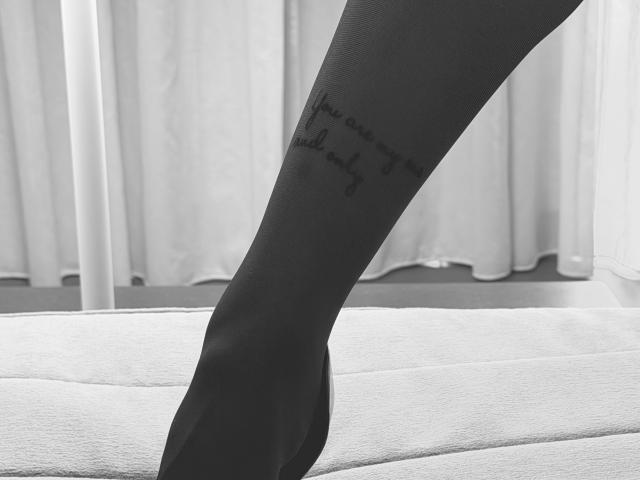 Le pied de BadAngel, vêtu de collants noirs, révèle un tatouage calligraphique sur la cheville interne, debout sur un tapis blanc texturé. L'image est en noir et blanc..